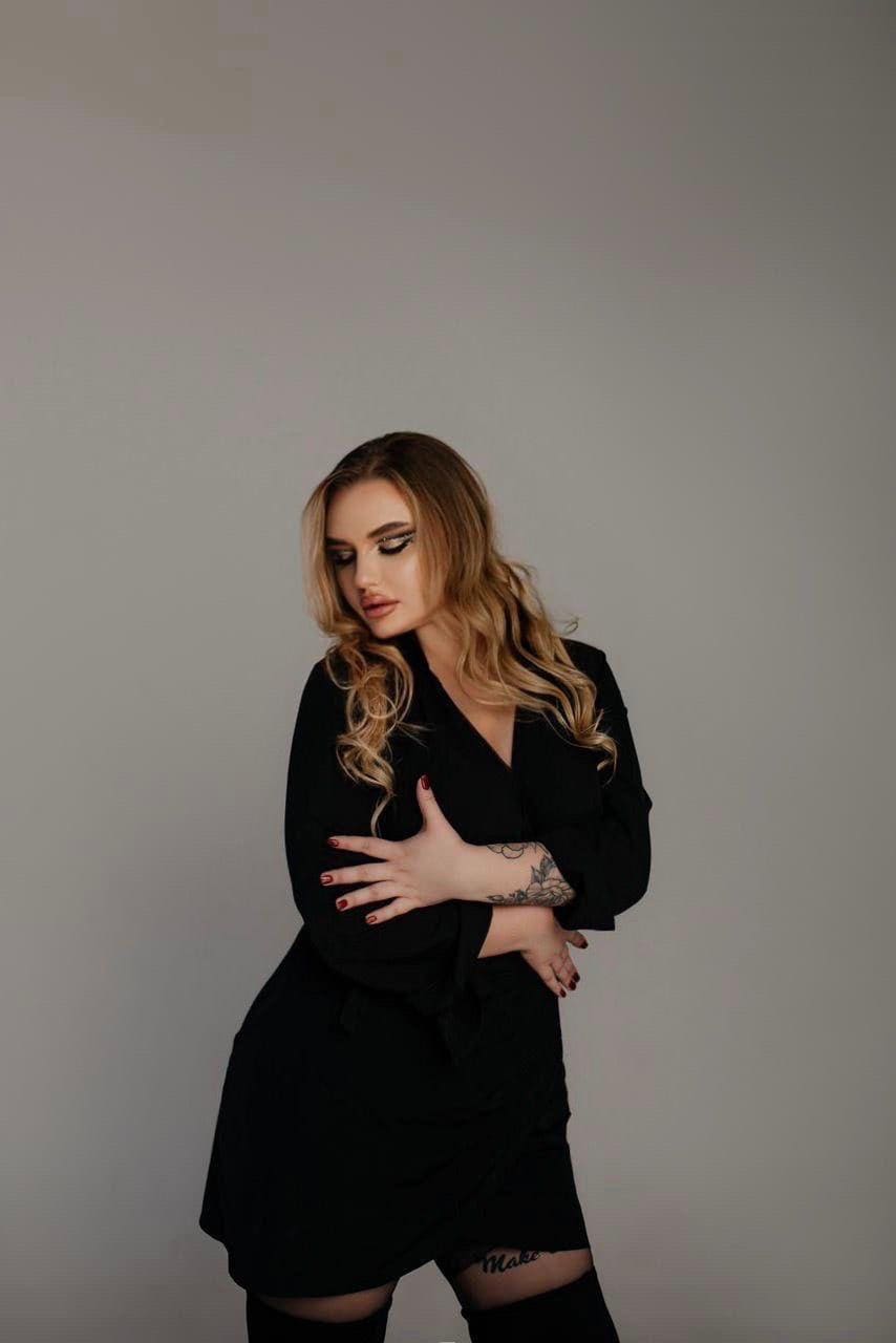 Katarina stands in a black wrap dress and thigh-high stockings against a gray backdrop, arms crossed, with tattoos visible on her forearm and thigh.