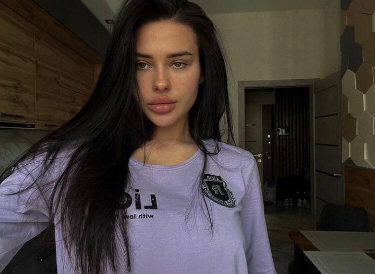 A woman stands indoors facing the camera, wearing a light purple long-sleeved shirt, near wooden cabinetry.