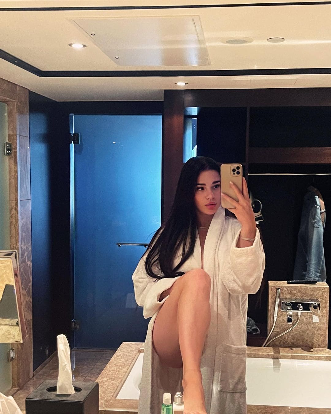 A woman sits on a bathtub edge in an open white bathrobe, taking a mirror selfie with a smartphone while showing one bare leg.