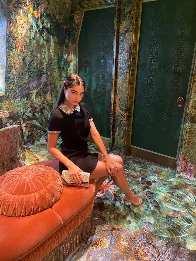 A woman sits on an orange velvet bench in a jungle-themed room, wearing a black dress with a jeweled collar.