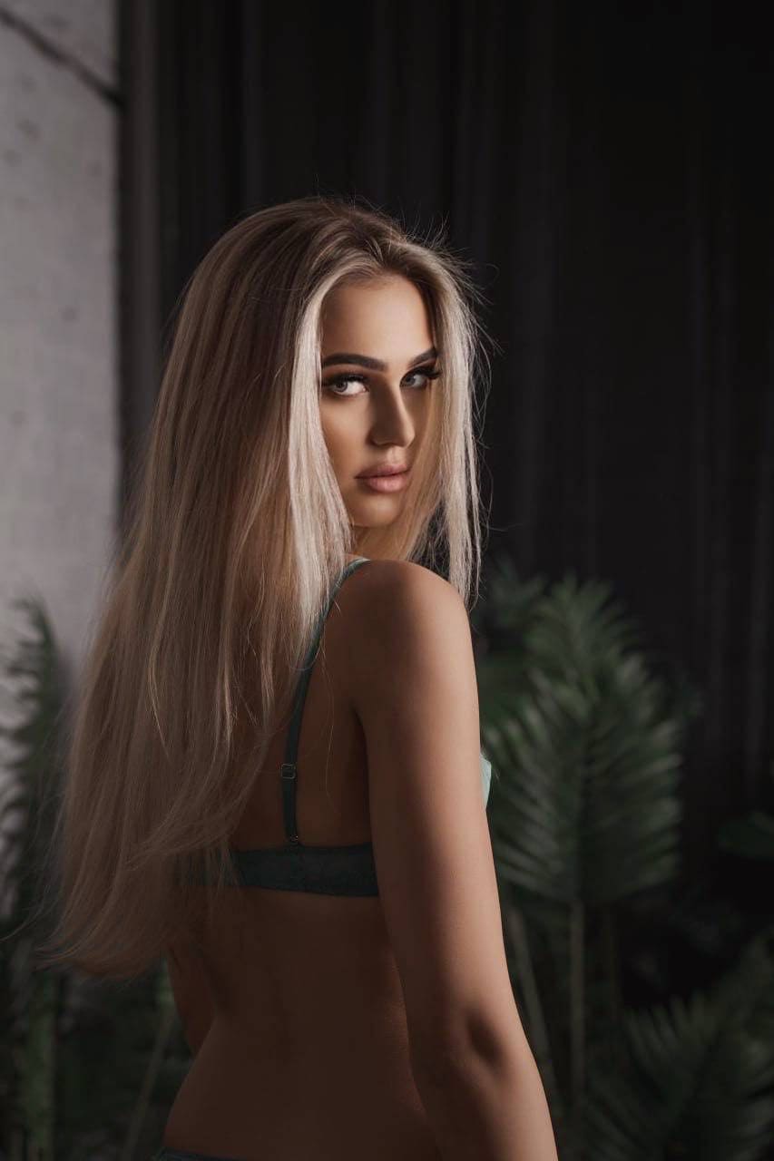 A woman in a dark green bra looks over her shoulder in profile, set against a dark background with blurred greenery.