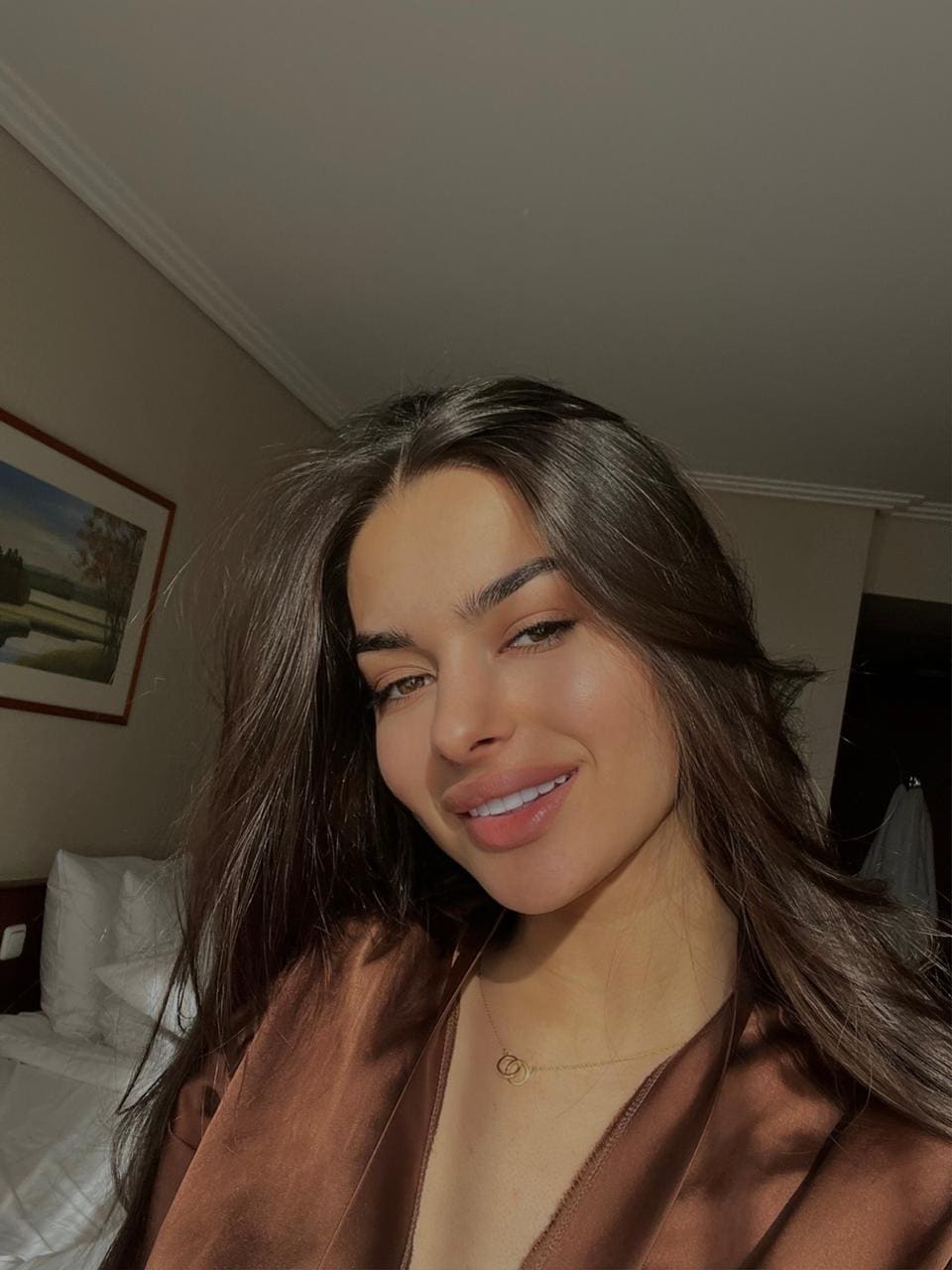 Tamila in a brown satin robe with a deep V-neckline, taking a selfie in a bedroom with a bed and framed landscape painting behind her.