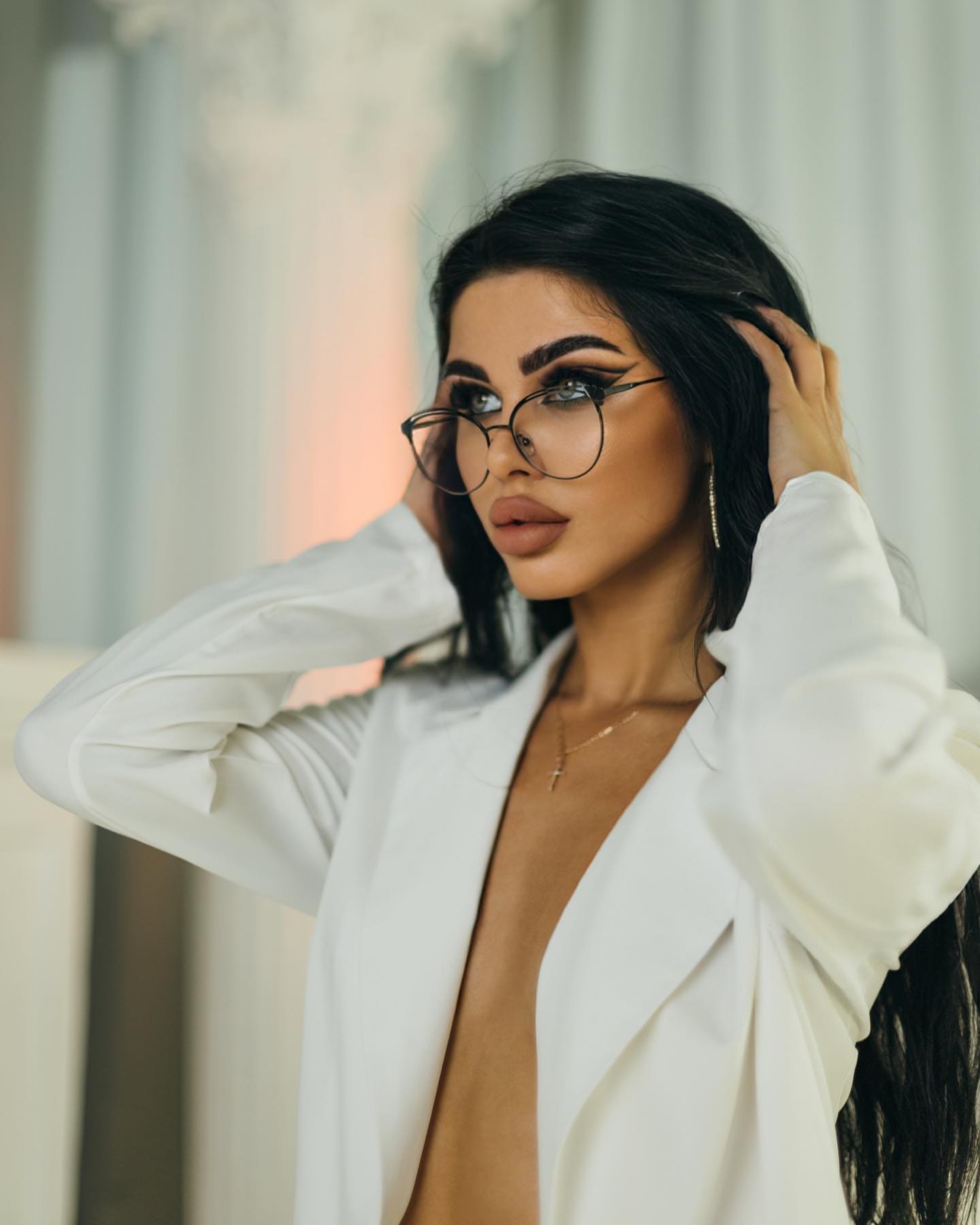 Chidem with long dark hair and round glasses, hands raised to head, wearing an open white blazer in a softly lit interior with pale columns.