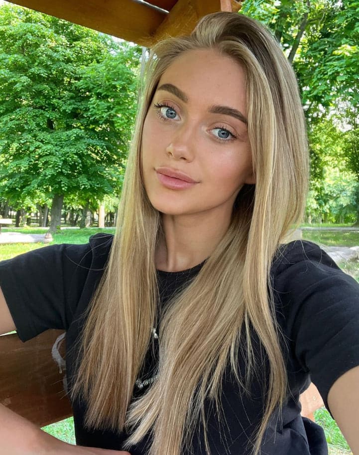 Nicole, with long blonde hair, wears a black t-shirt while seated outdoors under a wooden structure with green trees behind her.