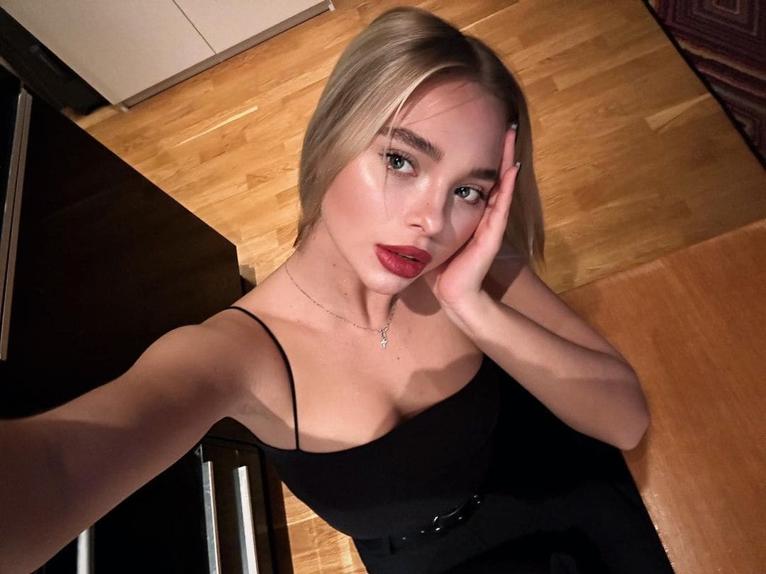 High-angle selfie of a blonde woman in a black spaghetti-strap top and thin necklace, resting her hand on her cheek in a room with wooden floors.