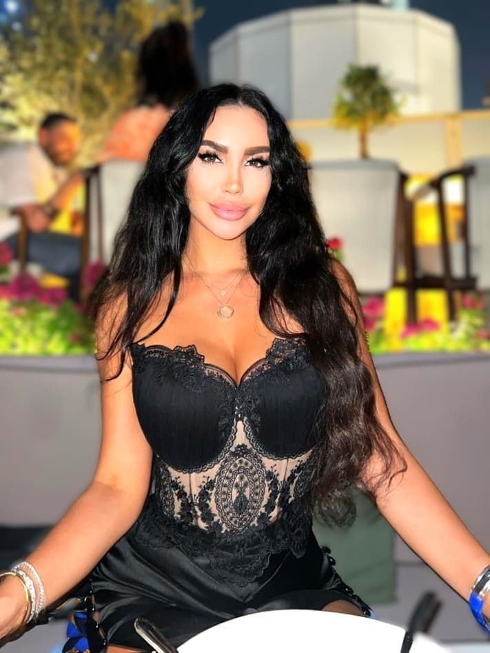 Alisa sits at an outdoor table in a black lace bodysuit with a deep neckline, wearing a pendant necklace and bracelets against a blurred background.