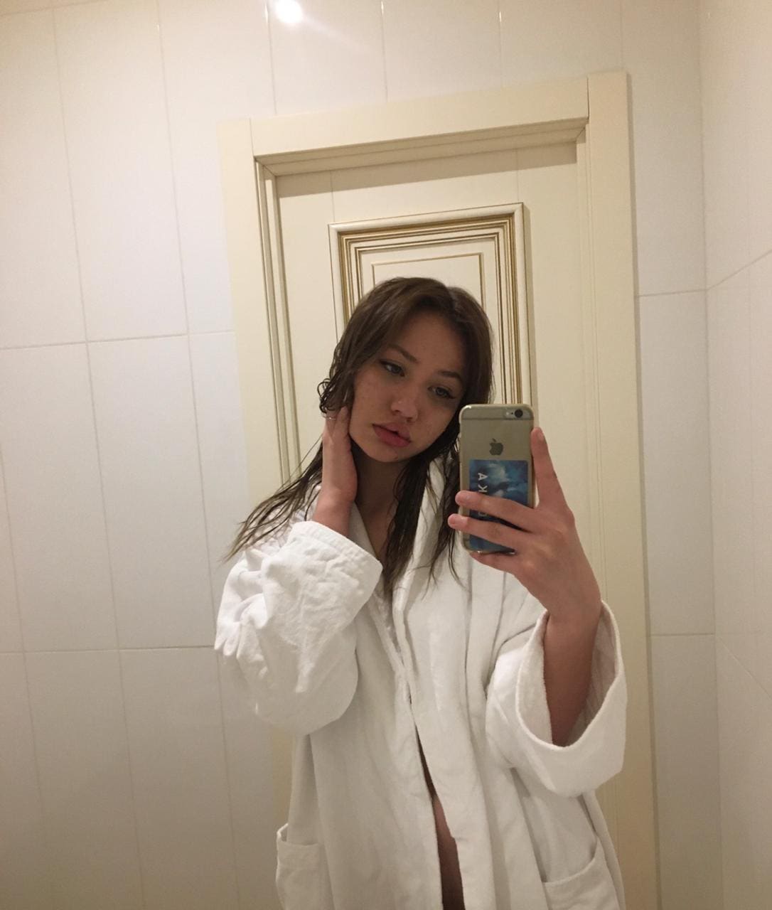 A woman with wet hair wears an open white bathrobe in a tiled bathroom, resting a hand on her neck while taking a mirror selfie.