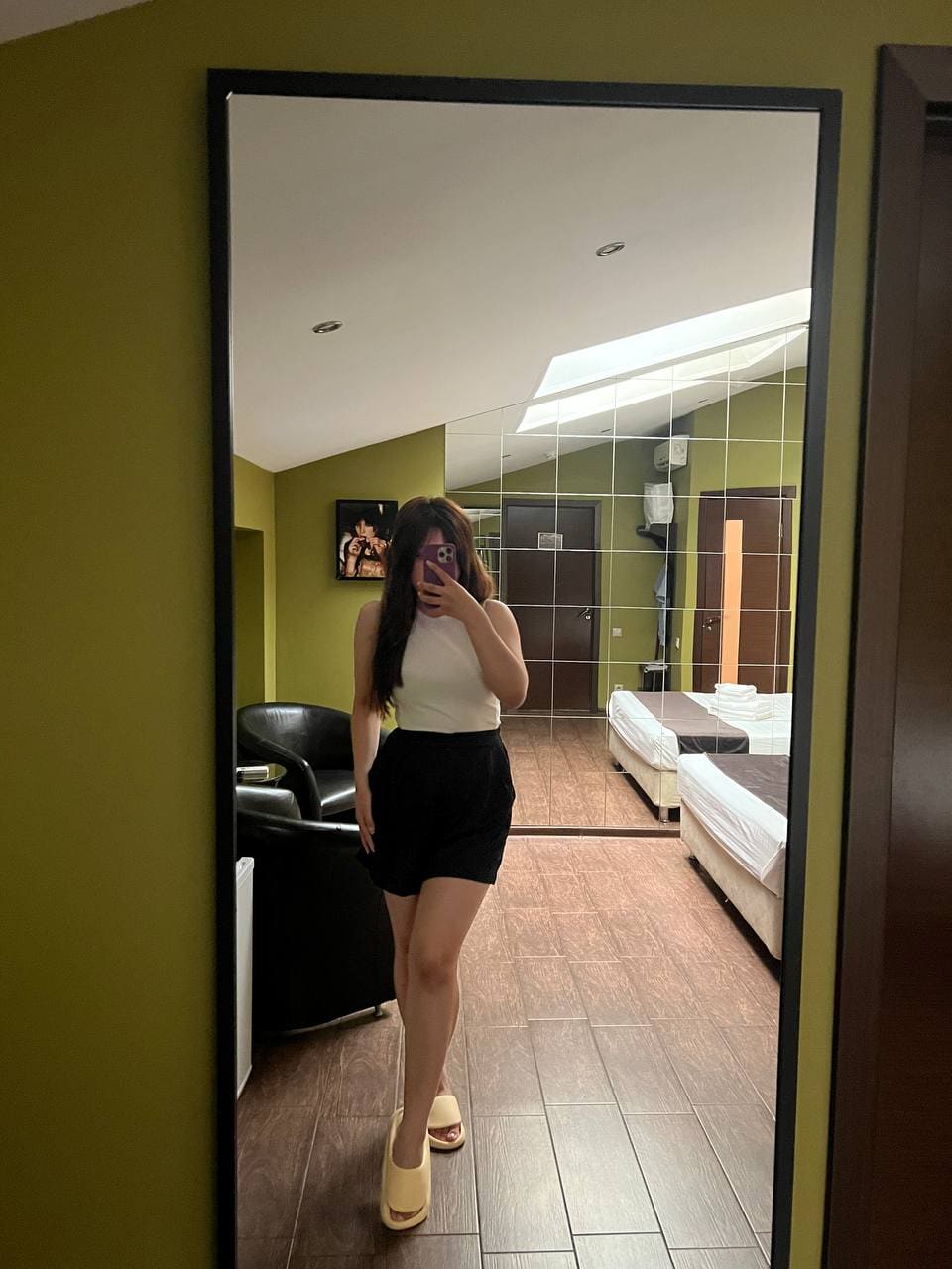 Standing before a full-length mirror against green walls, a woman wears a white top, black skirt, and yellow slip-on shoes.