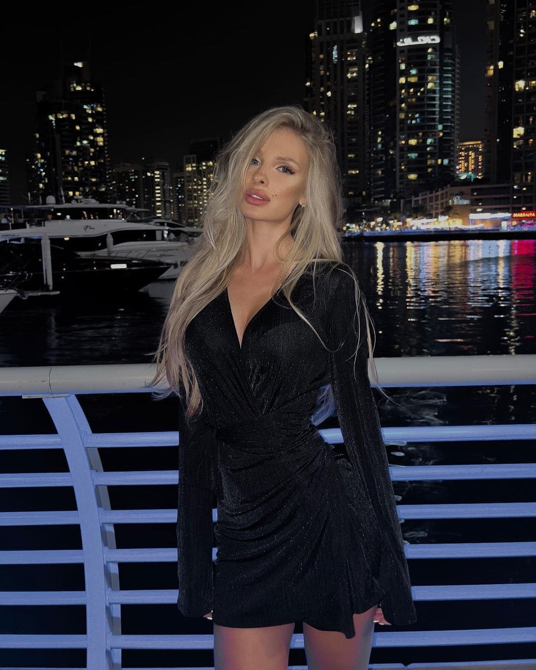 A woman in a black mini dress with a deep V-neck stands by a white railing at night, overlooking a city skyline and boats.