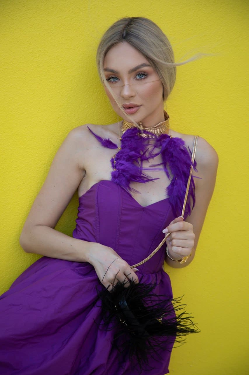 Bianca stands against a yellow wall in a strapless purple dress with a feathered neckline, holding a black feathered clutch and wearing a gold choker.