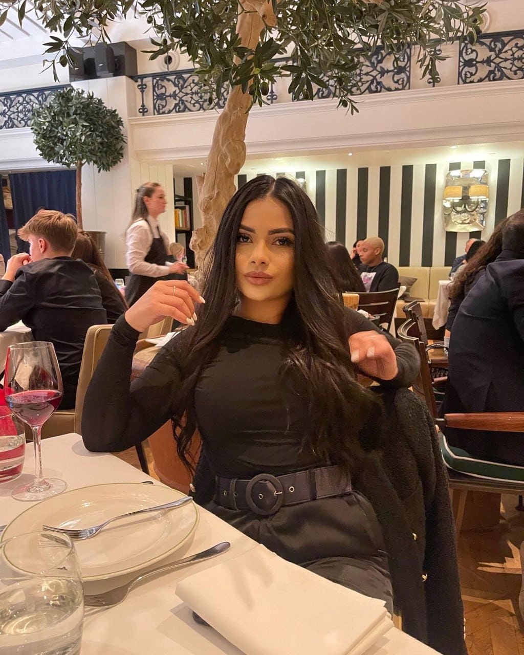 Angel sits at a restaurant table in a black long-sleeved top and wide belt, with red wine and a plate in front of her.