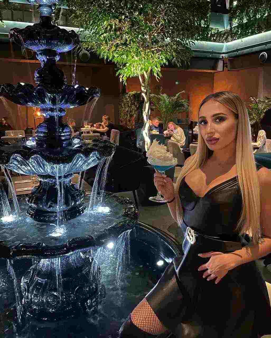 A blonde woman in a black dress and fishnets stands by an illuminated fountain holding a cocktail glass.
