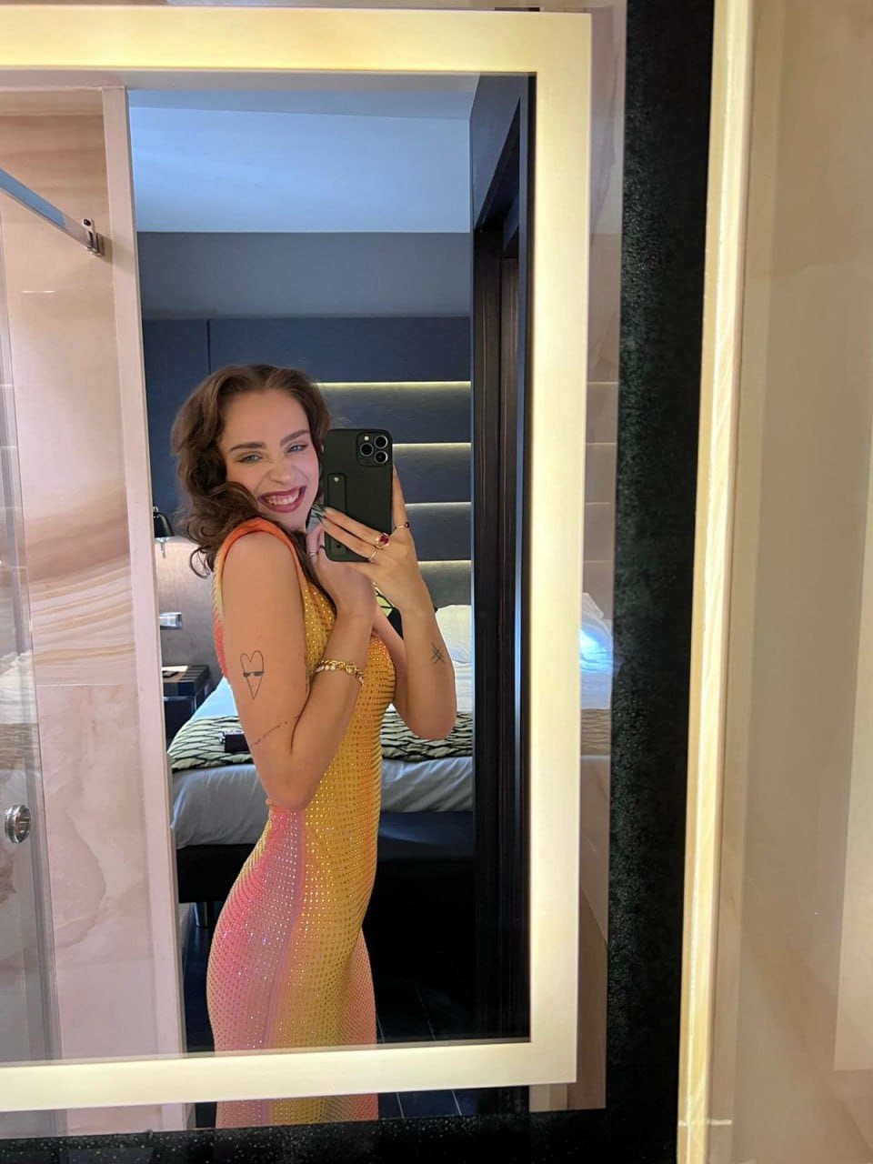 Mirror selfie in a bedroom showing a sleeveless yellow and pink gradient dress, phone held over the face.