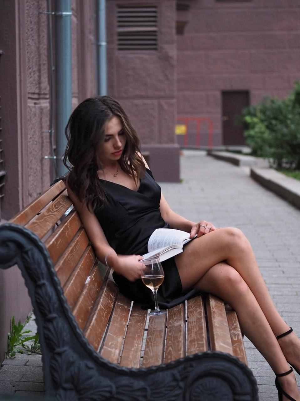 Sitting on a wooden bench in a short black dress and heels, holding an open book and a glass of white wine.