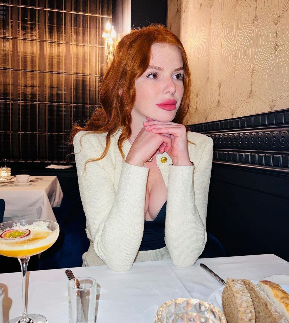 A red-haired woman sits at a restaurant table in a cream cardigan and dark top, hands clasped under her chin, with a cocktail and bread nearby.
