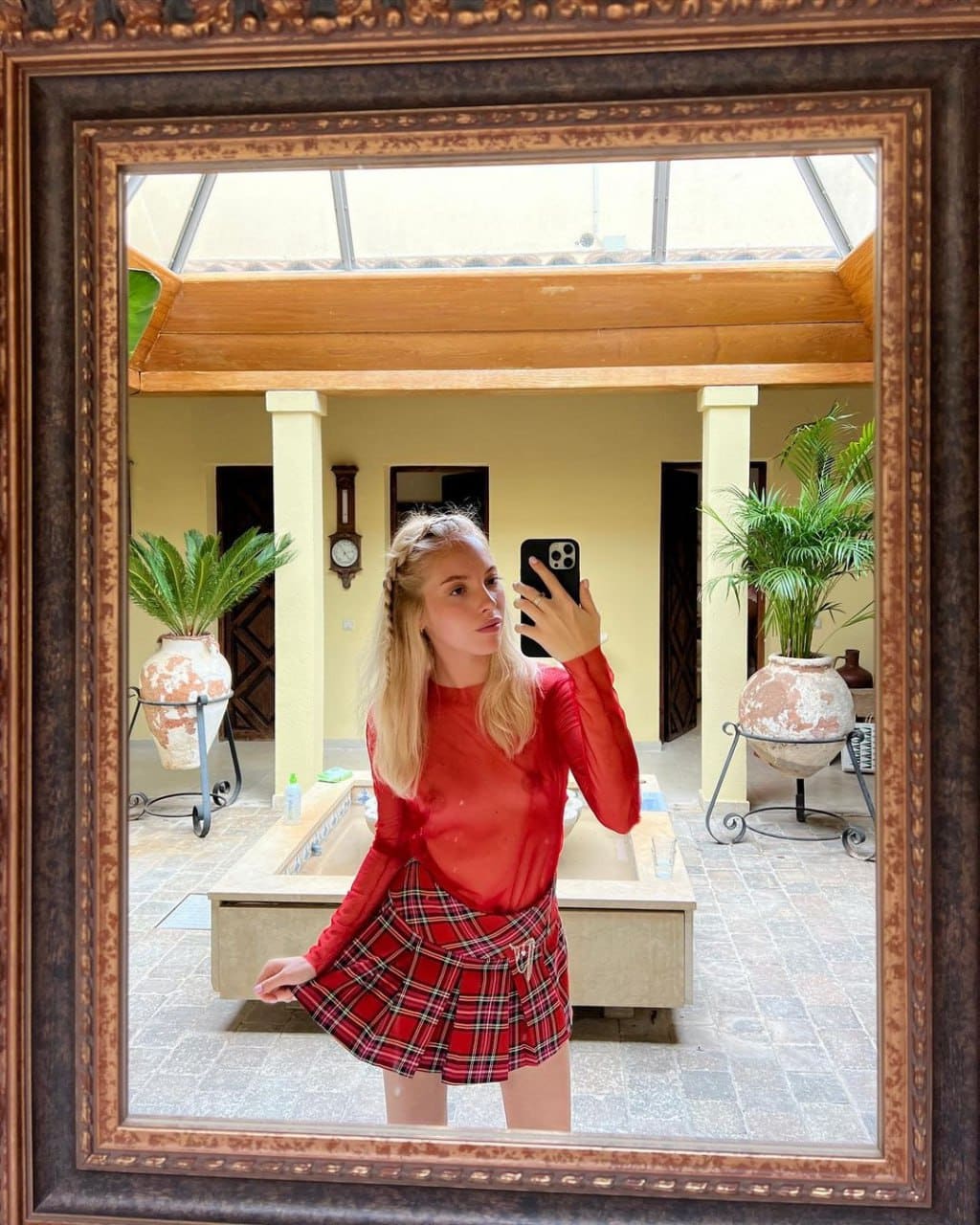 A blonde woman with a side braid takes a mirror selfie in a red top and plaid skirt, standing in a tiled courtyard.