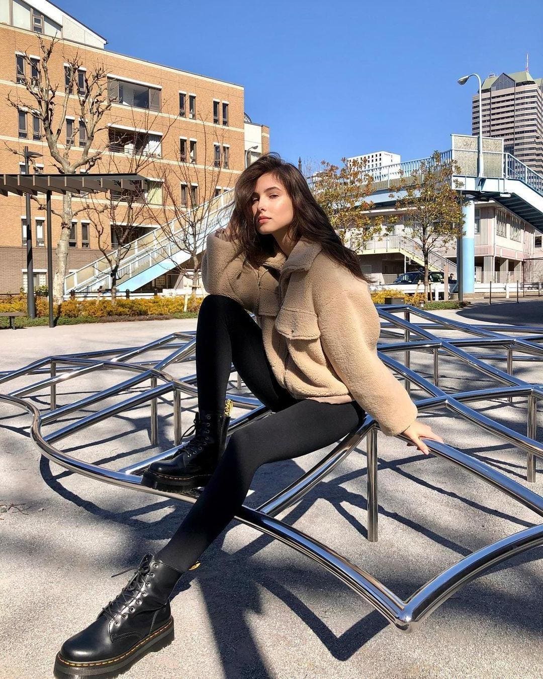 A woman sits on a curved metal sculpture in an outdoor plaza, wearing a beige fleece jacket, black leggings, and lace-up boots.