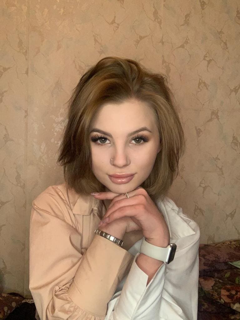Ruslana rests her chin on clasped hands, wearing a pink blouse under an open white shirt against a textured beige wall.
