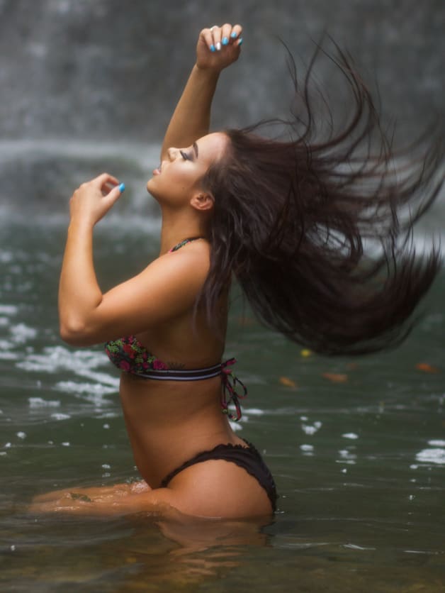 A woman stands waist-deep in water in a floral bikini, one arm raised, head tilted back as wind blows her hair sideways.
