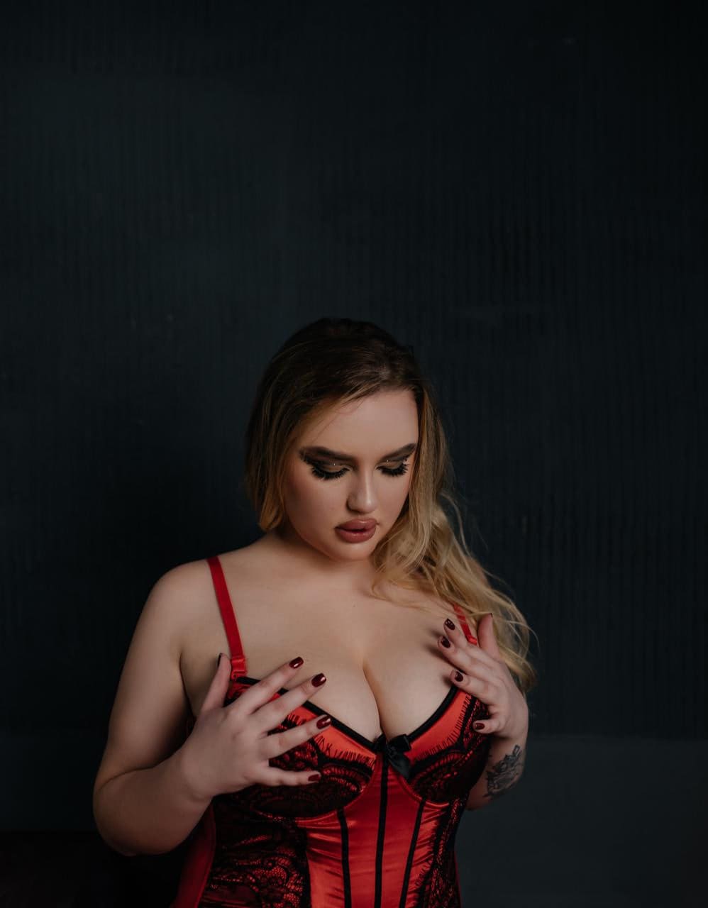 A woman with dark eye makeup wears a red and black lace corset against a dark background, her hands resting on the front of the garment.