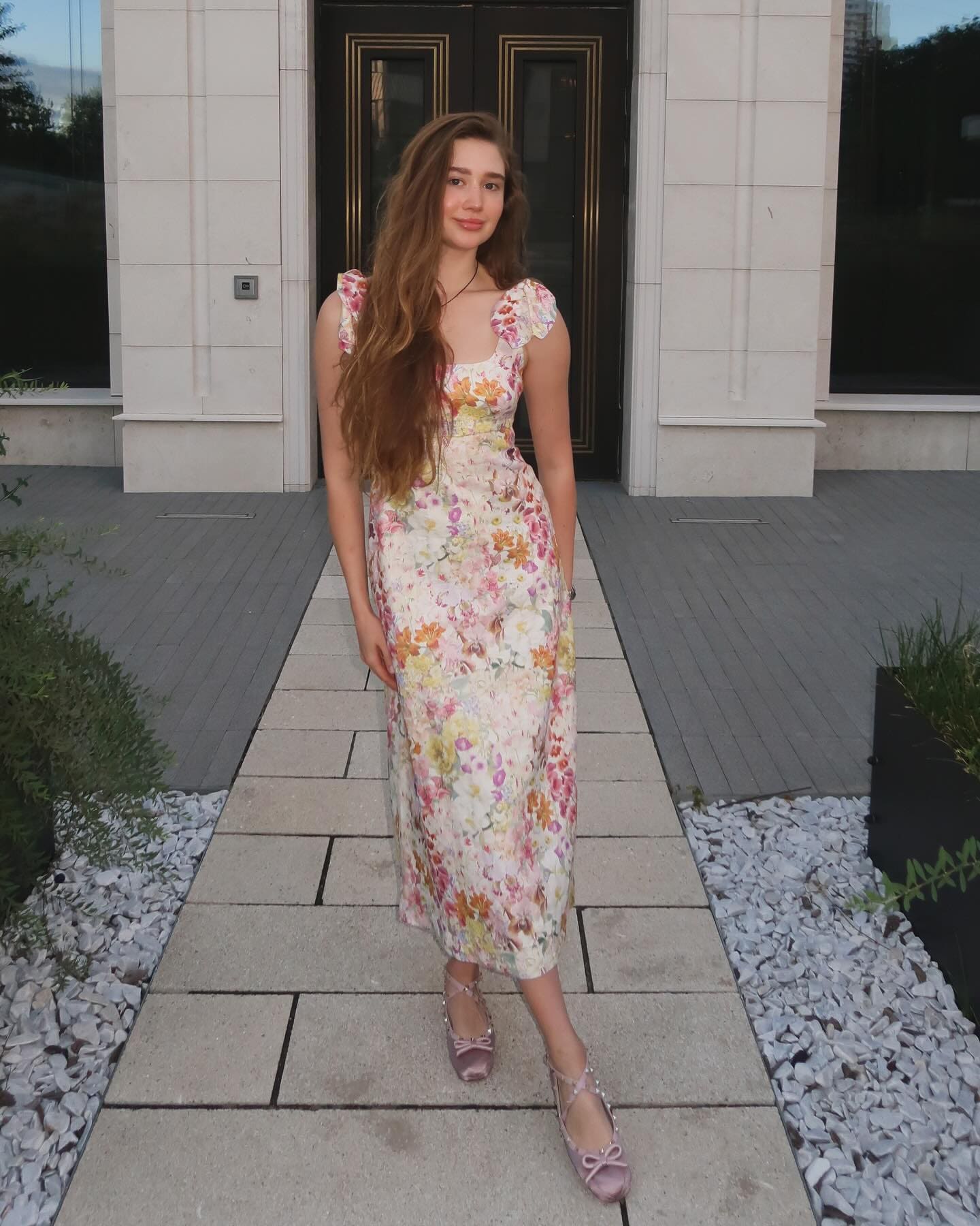 Aeneas stands on a stone walkway wearing a floral midi dress with ruffled sleeves and pink flat shoes.