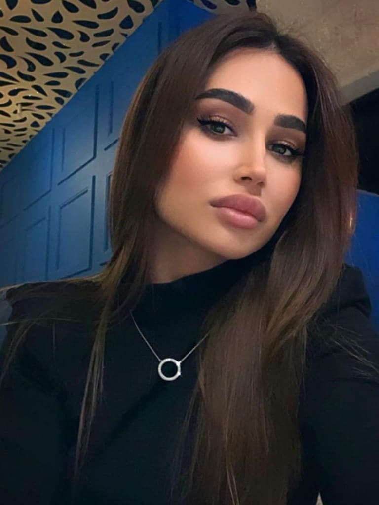 Amina wears a black high-neck top and silver necklace, posing before a blue paneled wall and patterned ceiling.