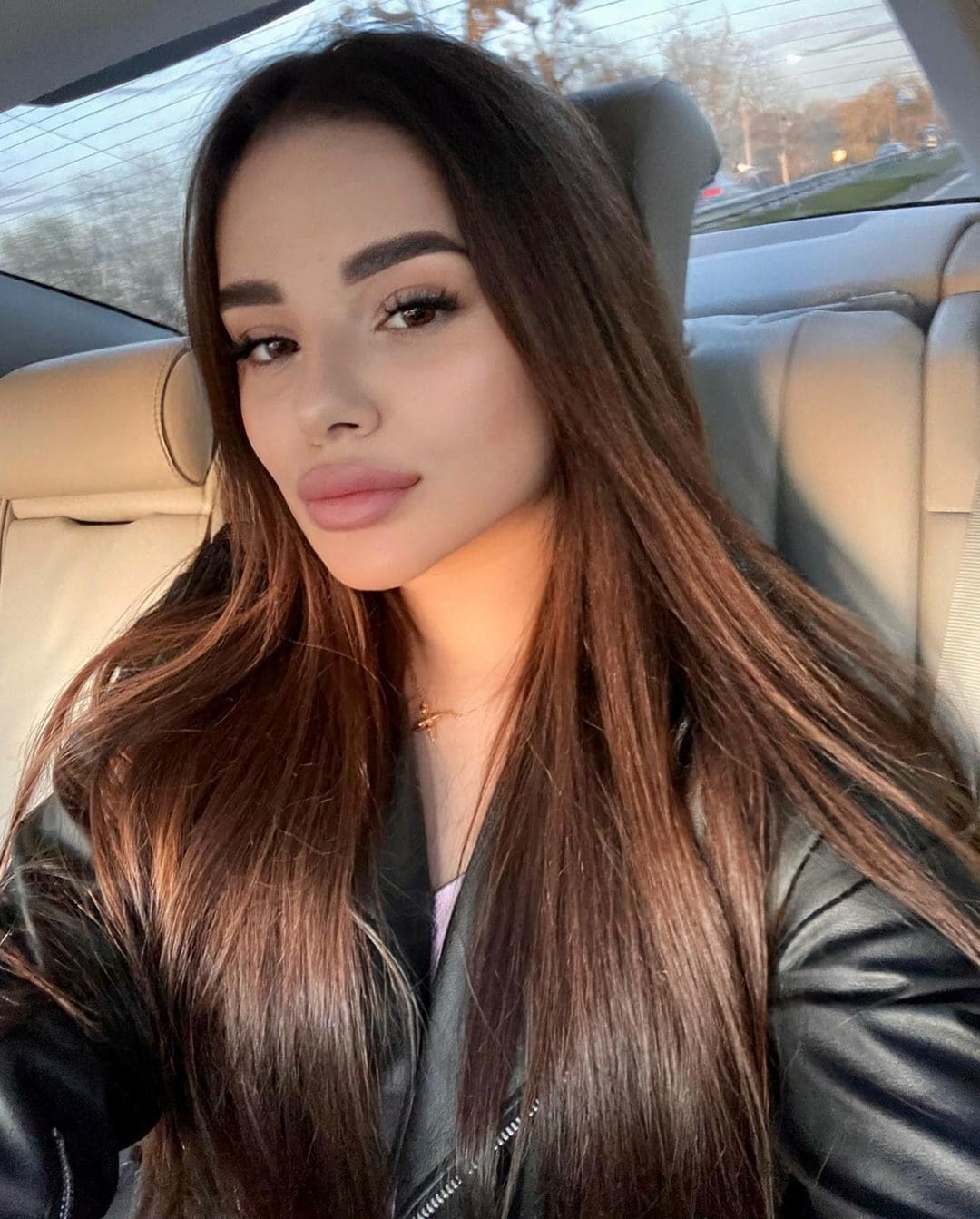 A woman with straight brown hair sits in a car back seat wearing a black leather jacket over a light top, with a thin gold necklace.