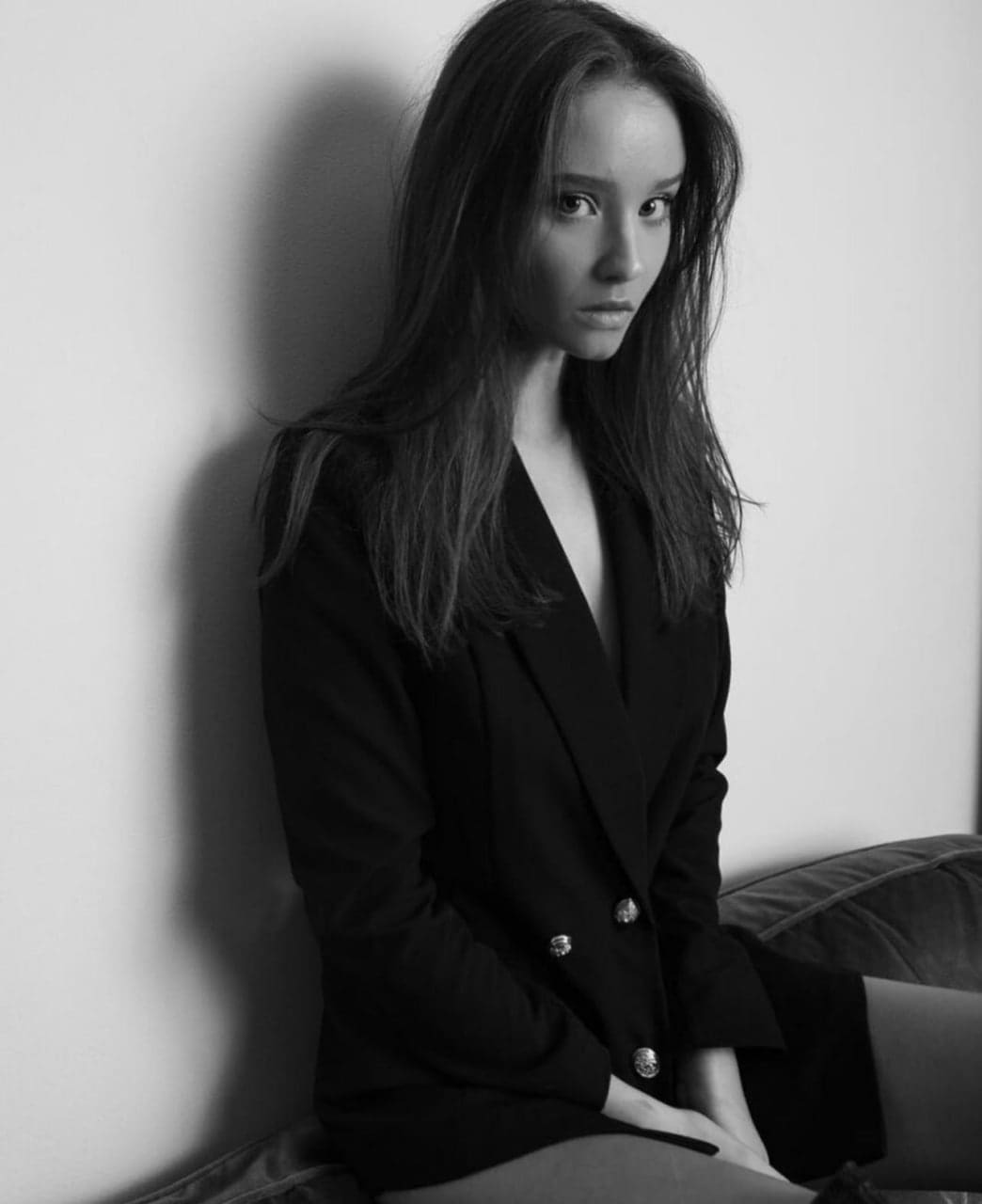 Seated on a couch wearing an open black double-breasted blazer, hands resting in lap against a plain wall.