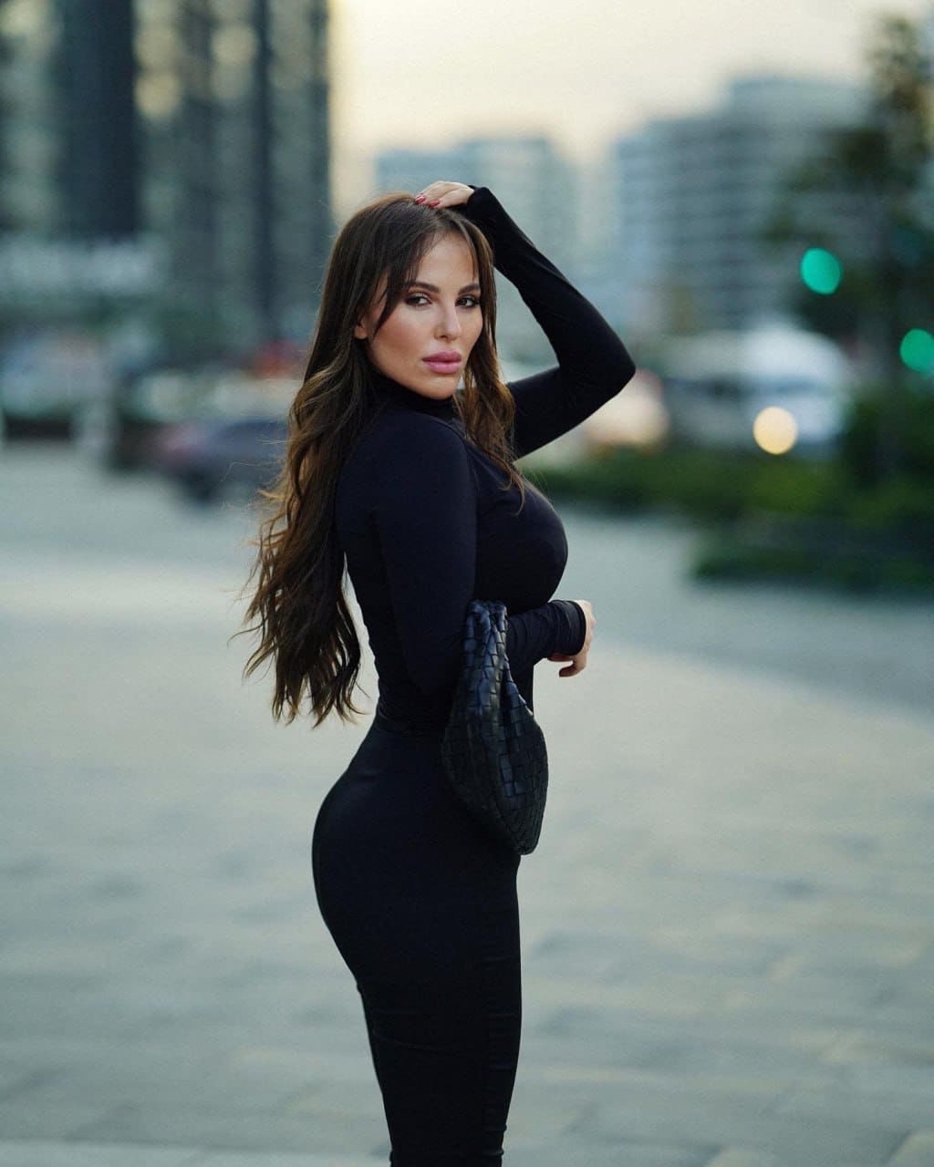 A woman in profile wears a black long-sleeve turtleneck dress, holding a small woven handbag at her hip with one hand on her head against blurred city buildings.