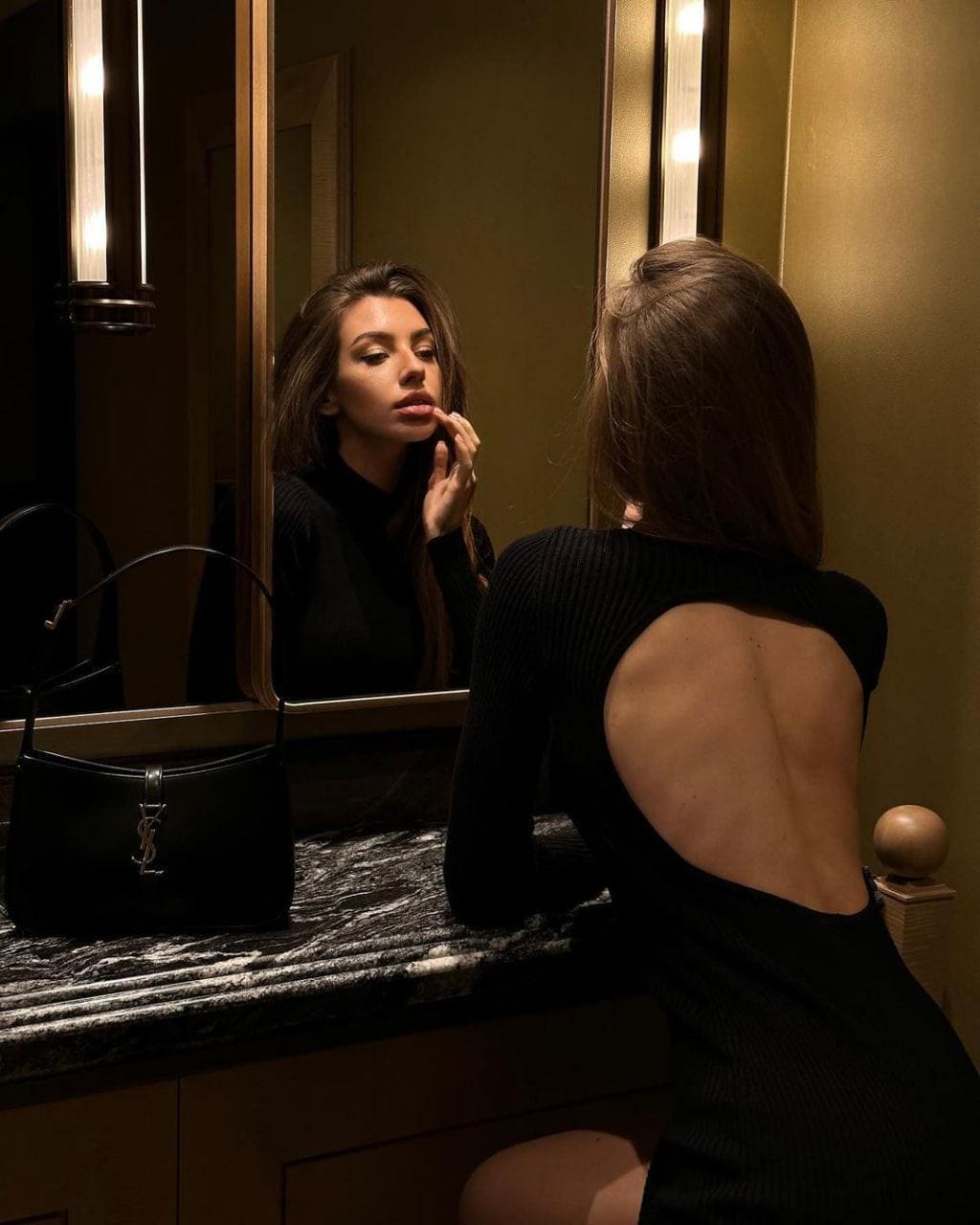 Seen from behind, a woman sits at a marble vanity in a black ribbed top with an open back, her reflection touching her lips in the mirror.