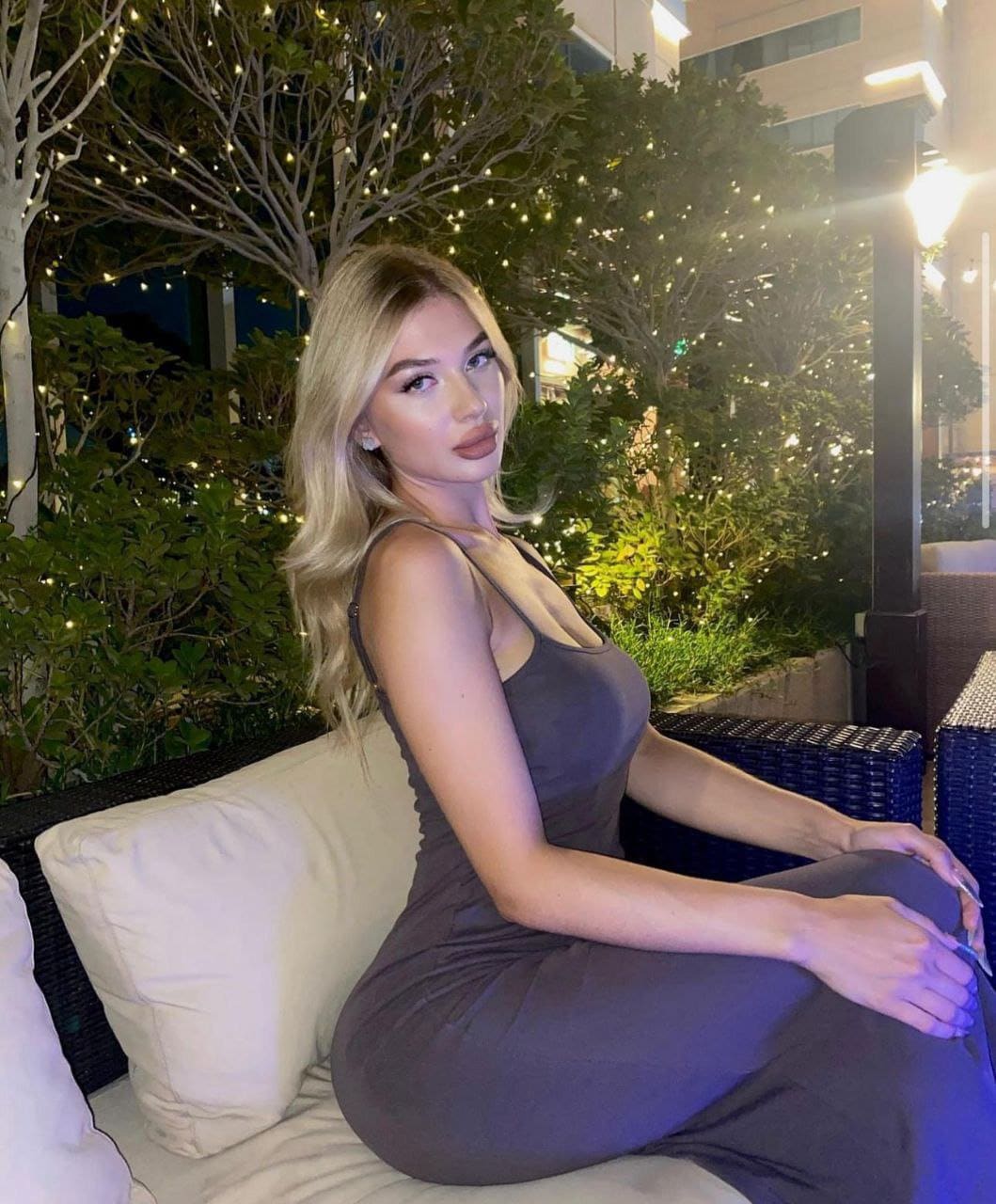 A blonde woman sits on an outdoor sofa in a dark grey sleeveless dress, with leafy bushes and string lights behind her at night.