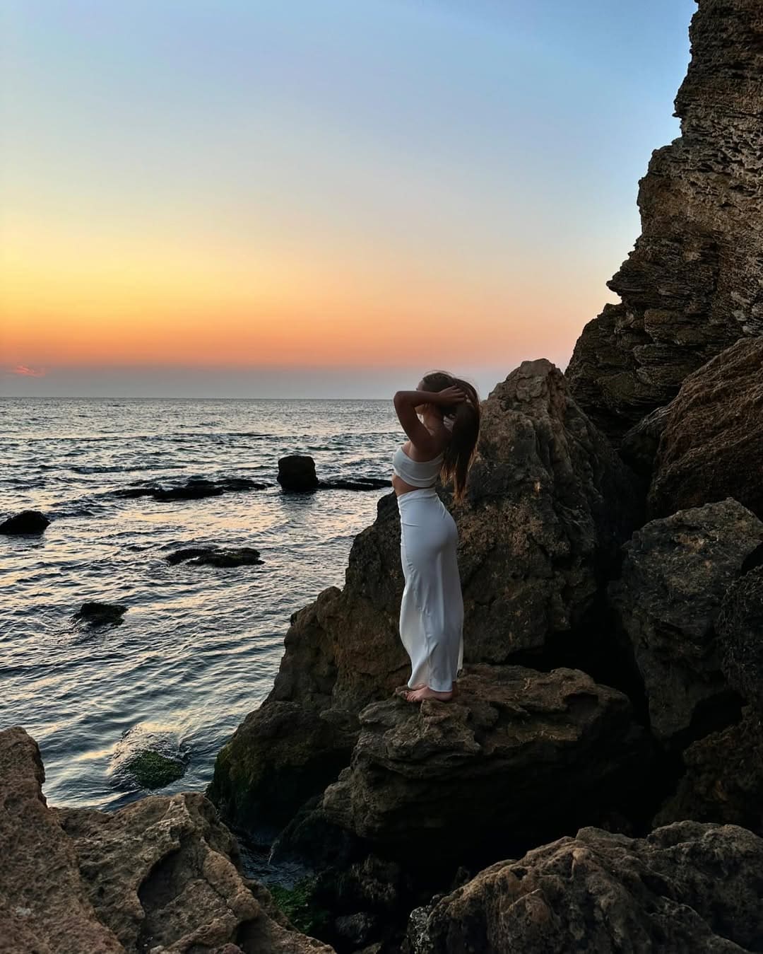 A woman in a white crop top and long skirt stands barefoot on a rocky sea outcrop at sunset, with both hands raised to her head.