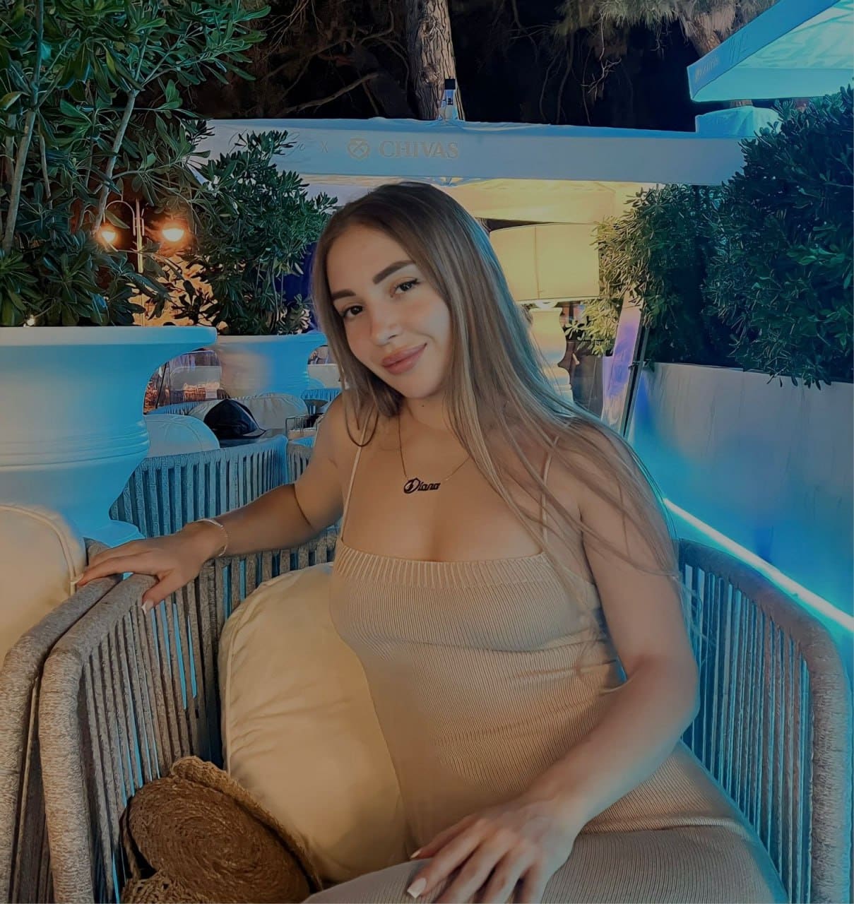 A woman sits in a woven chair at night, wearing a beige ribbed dress and necklace, surrounded by potted plants and blue patio lighting.