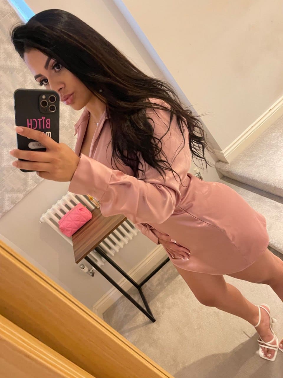 A mirror selfie shows a woman in a pink satin mini dress and white heels, standing near a radiator and small table.