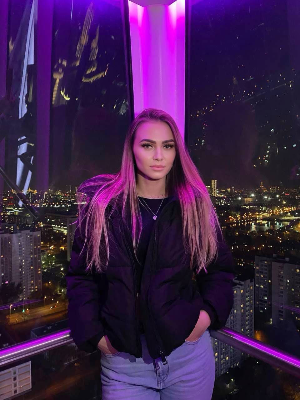 A woman in a black puffer jacket and jeans stands on a balcony, hands in pockets, against a purple-lit cityscape.