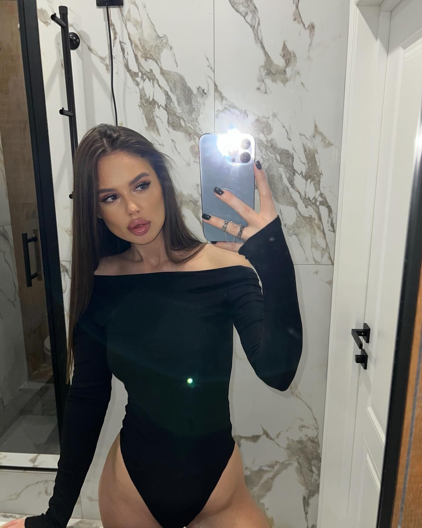 A woman with long dark hair takes a mirror selfie in a marble bathroom, wearing a black off-the-shoulder bodysuit.