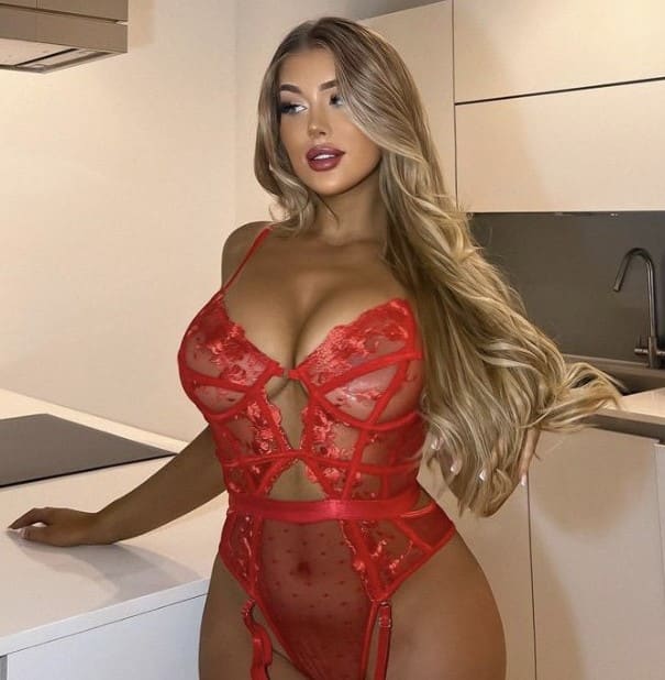 A blonde woman with wavy hair stands in a modern kitchen wearing a red lace and mesh bodysuit with cutouts, resting one hand on a white countertop.
