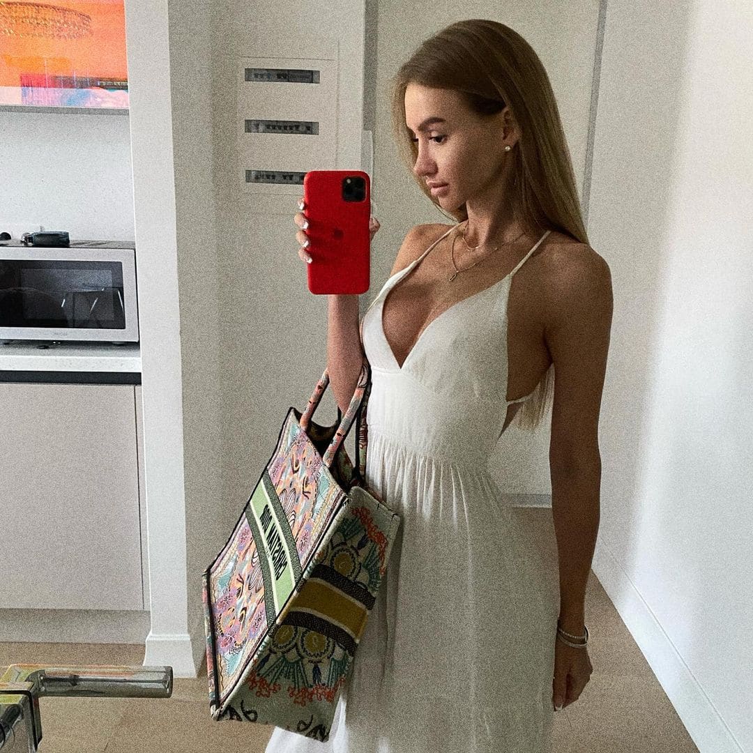 A woman in a white sleeveless V-neck dress takes a mirror selfie with a red phone, holding a large patterned tote bag in a white room.