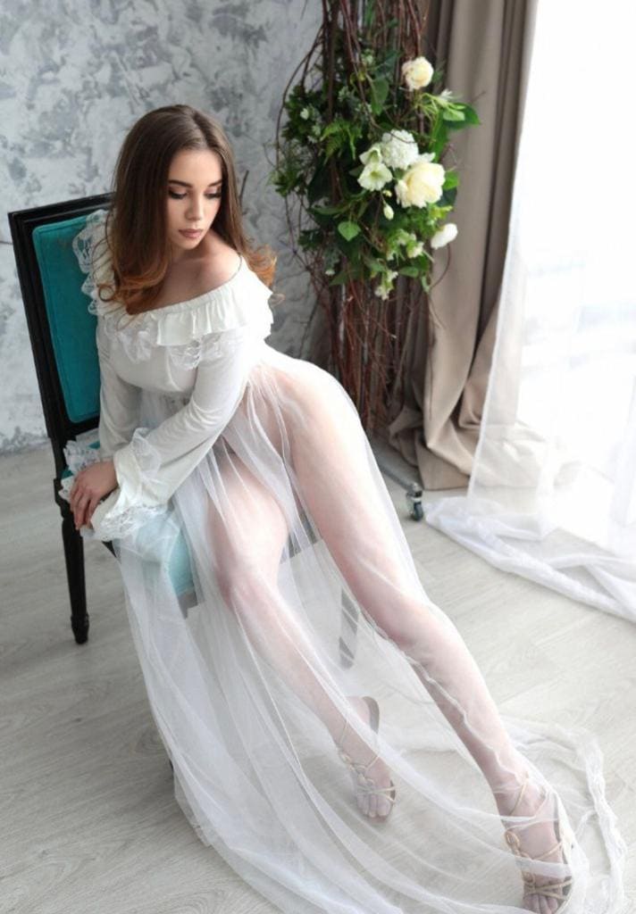 Seated on a teal chair, she wears a sheer white dress with ruffled sleeves and a high slit, with a window and flowers in the room.