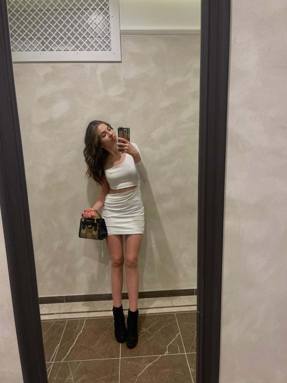 Mirror selfie in a tiled room showing a white one-shoulder crop top, matching skirt, black boots, and a patterned handbag.