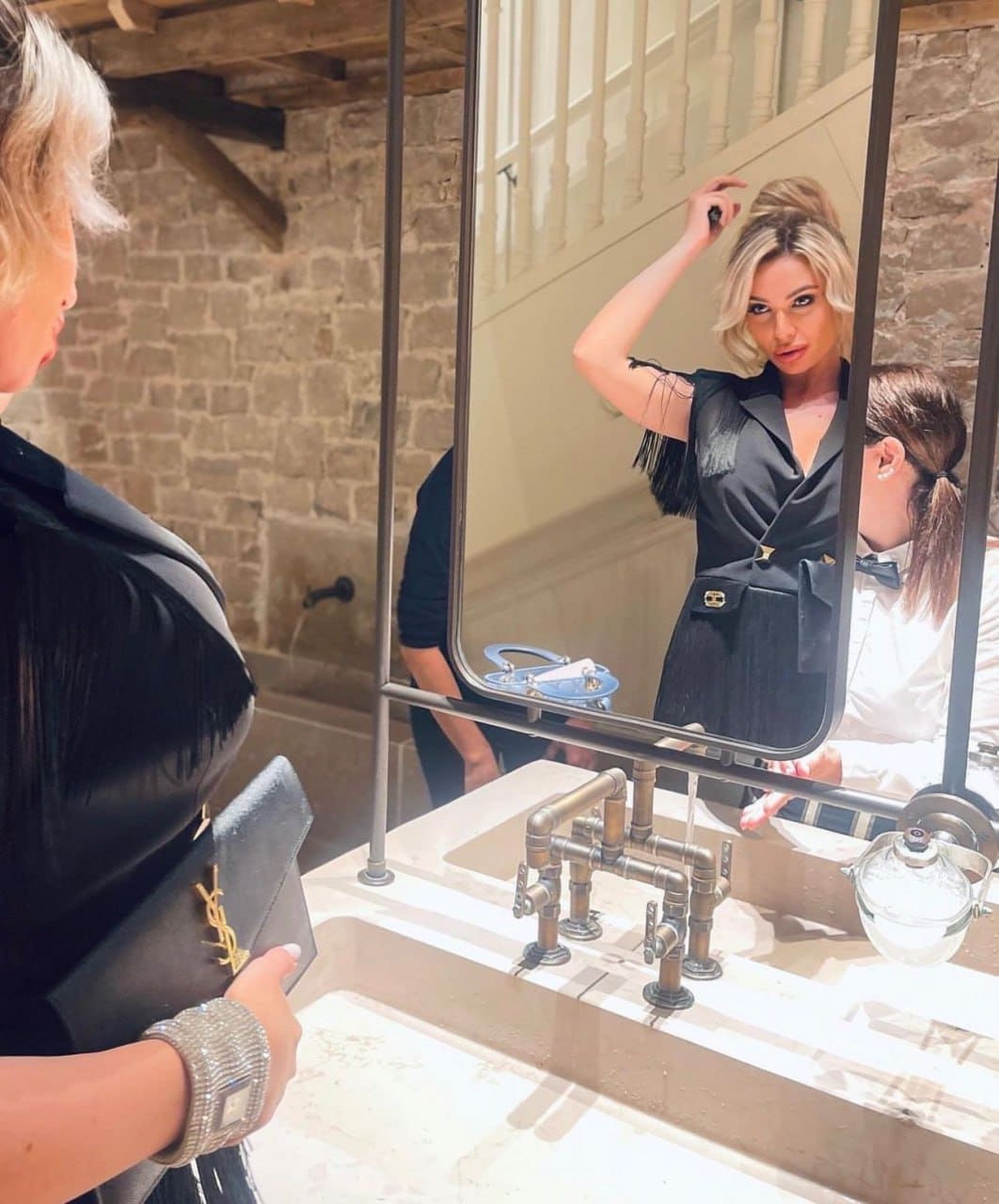 A woman in a black fringed dress stands before a mirror in a stone room, holding a clutch and touching her updo.