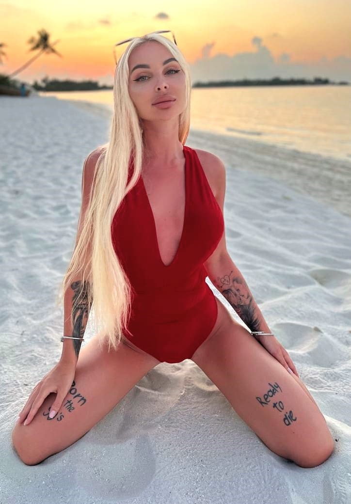 A blonde woman kneels on a sandy beach at sunset wearing a red deep-V swimsuit, with sunglasses on her head and visible tattoos.