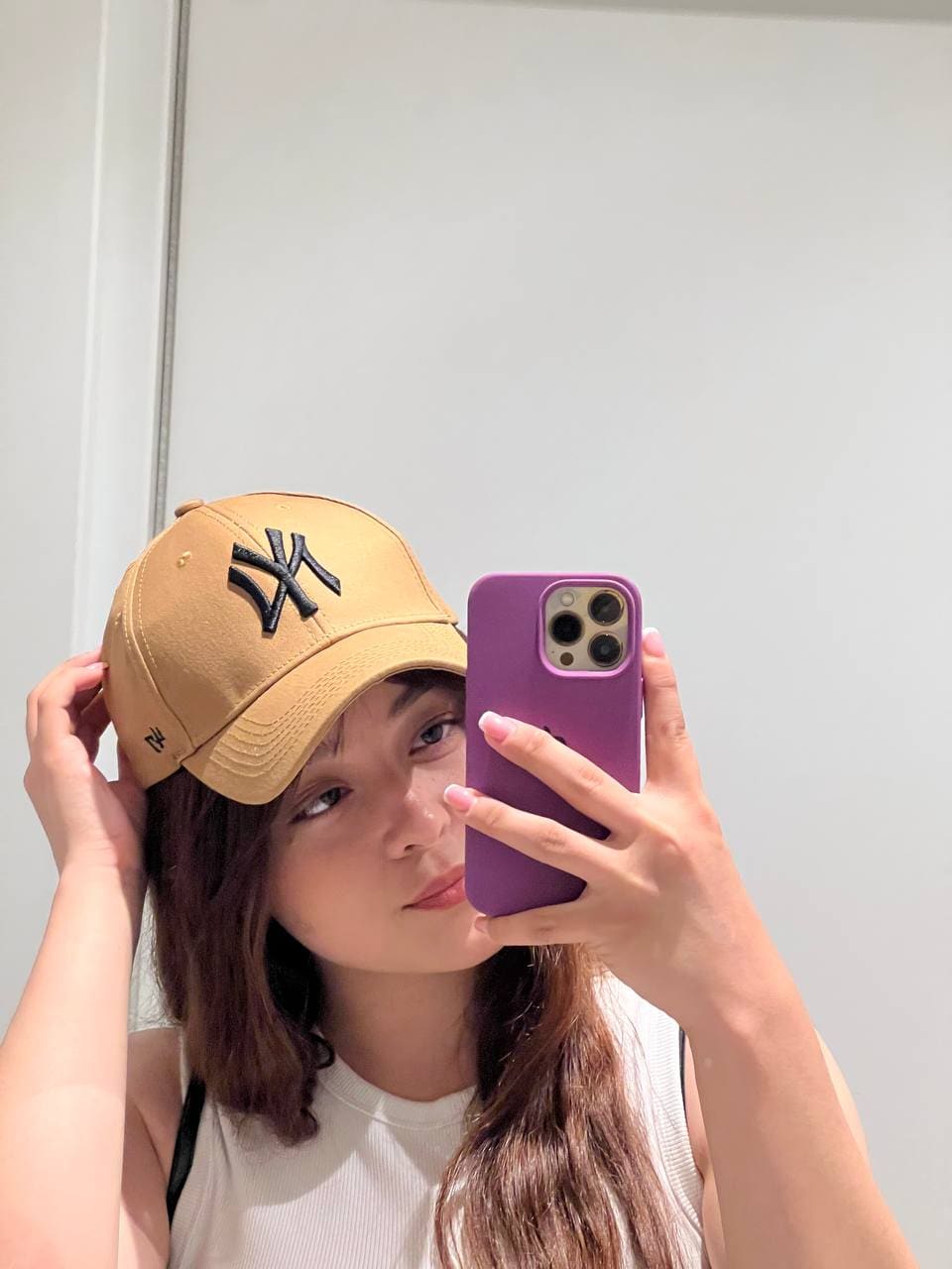 Leyla takes a mirror selfie in a tan cap and white sleeveless top, holding a purple phone against a plain white wall.