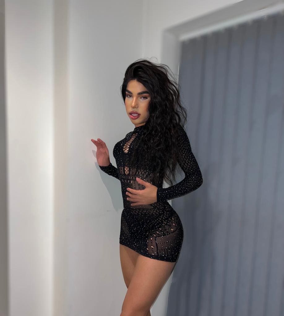 Sofa stands in a white room wearing a short black dress with sheer mesh and studs, one hand on her hip and the other raised near the wall.