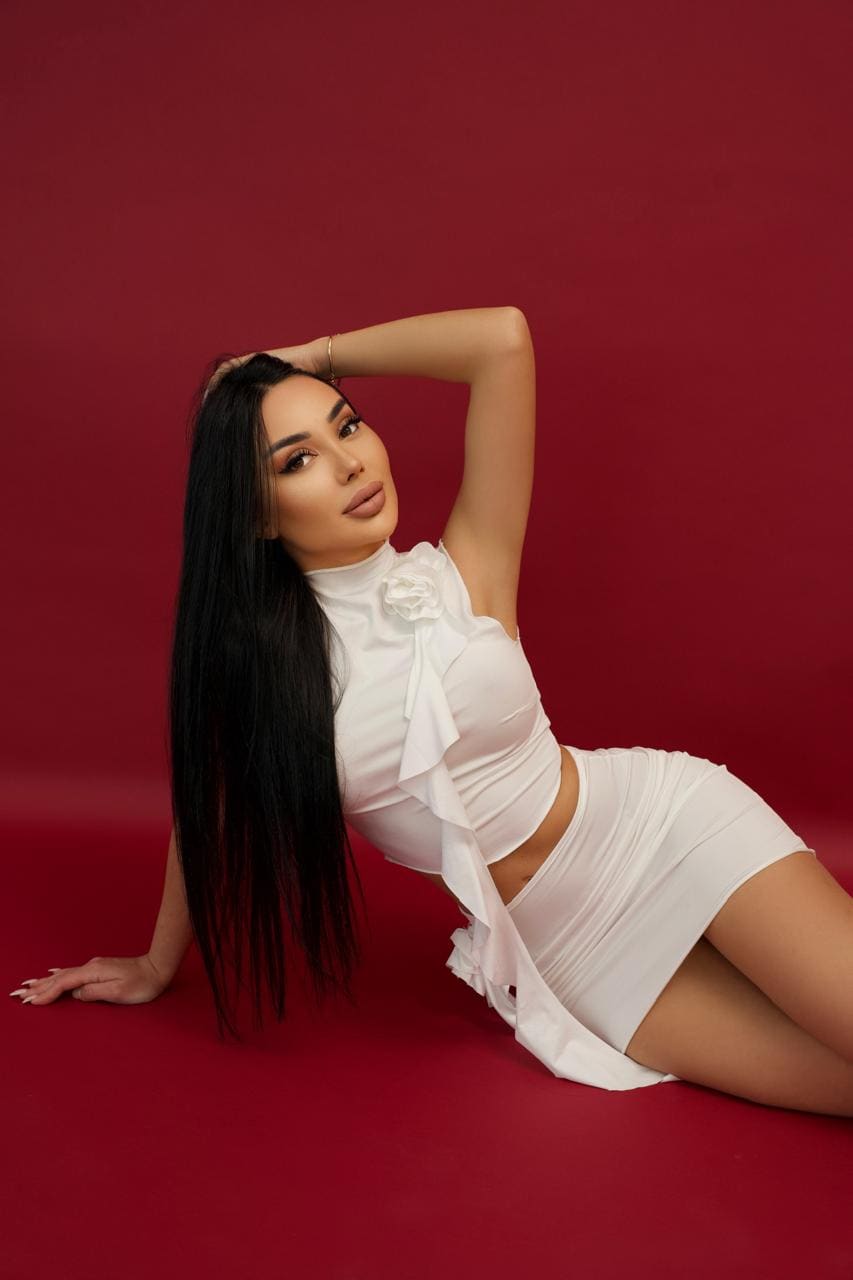 A woman lies on a red floor in a white high-neck ruffled crop top and skirt, one arm raised behind her head, the other on the floor.
