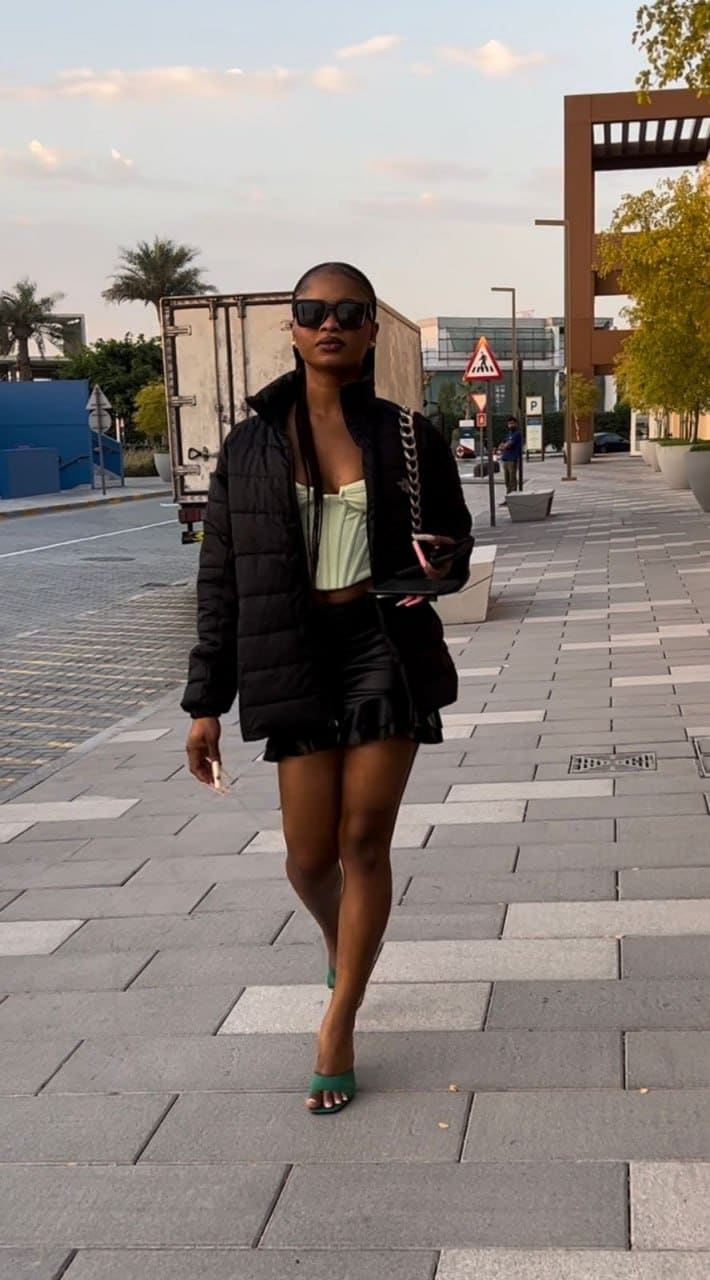 A woman with a shaved head and sunglasses walks a city sidewalk in a black puffer jacket, green top, short skirt, and green heels, holding a phone.