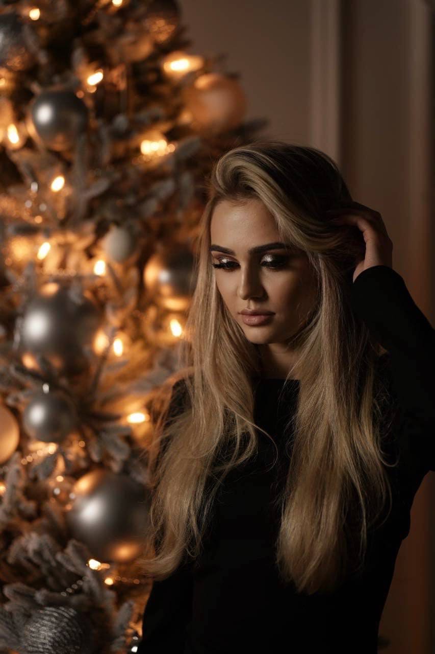 A woman in a black long-sleeved top stands before a lit Christmas tree, touching her hair with one hand.