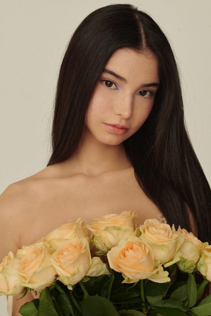Sabrina holds a bouquet of pale peach roses against her bare shoulders and chest against a plain light background.