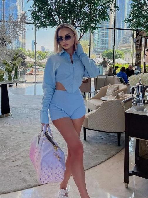 A blonde woman in a light blue cut-out shirt and shorts stands in a sunlit lounge with large windows, holding a patterned bag and wearing sunglasses.