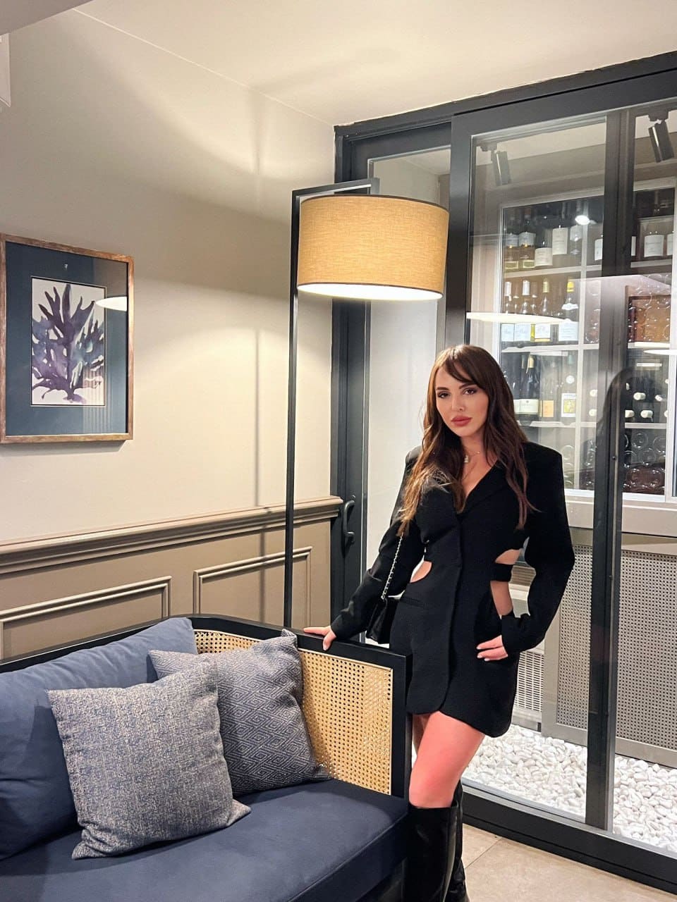 A woman in a black blazer dress with side cutouts and knee-high boots stands indoors near a blue sofa, with a floor lamp and glass cabinet behind her.